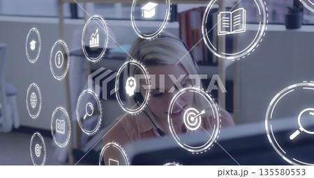 Focusing office worker wearing headset analyzing data on computer at desk, with digital icons 135580553