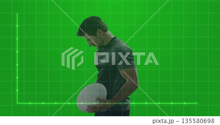 Posing male wearing dark sportswear holding white oval ball in green screen set, with grid backdrop Posing male wearing dark sportswear holding white oval ball in green screen set, with grid backdrop 135580698