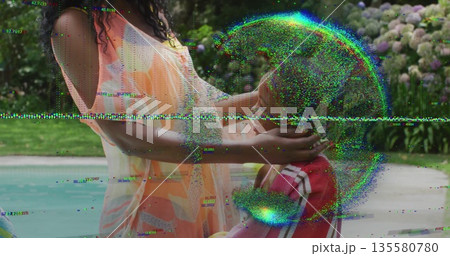 Adjusting mother placing straw sun hat on daughter in striped top by swimming pool, stone deck 135580780