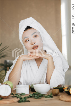 A young woman wearing a white bathrobe and towel on her head applies a sheet mask for skincare while surrounded by fresh fruits and natural ingredients 135581221