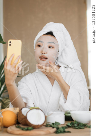 A young woman with a towel on her head and a sheet mask on her face takes a selfie with her phone while wearing a white bathrobe A young woman with a towel on her head and a sheet mask on her face takes a selfie with her phone while wearing a white bathrobe 135581225