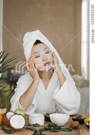 A woman with a towel on her head and wearing a bathrobe applies a facial sheet mask while surrounded by fruits and skincare products 135581230