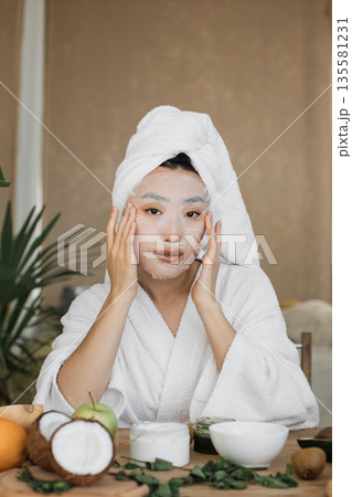 A young woman with a towel on her head and a white robe applies a hydrating sheet mask to her face while surrounded by fresh fruits and skincare products 135581231