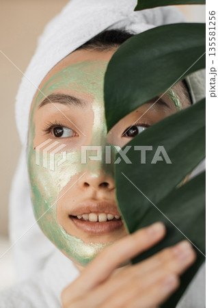 A young woman with a white towel wrapped around her head and a green face mask applied to her skin is partially hidden by a large green leaf A young woman with a white towel wrapped around her head and a green face mask applied to her skin is partially hidden by a large green leaf 135581256