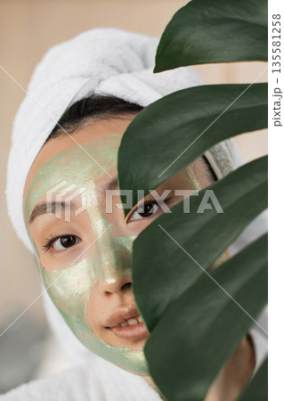 A young woman with a white towel wrapped around her head and a green face mask applied to her skin is partially obscured by a large green leaf, suggesting a natural beauty treatment A young woman with a white towel wrapped around her head and a green face mask applied to her skin is partially obscured by a large green leaf, suggesting a natural beauty treatment 135581258