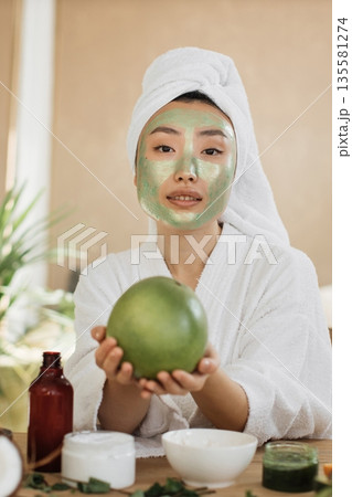 A young Asian woman with a white towel wrapped around her head and a green face mask on her face holds a green fruit while wearing a white robe A young Asian woman with a white towel wrapped around her head and a green face mask on her face holds a green fruit while wearing a white robe 135581274