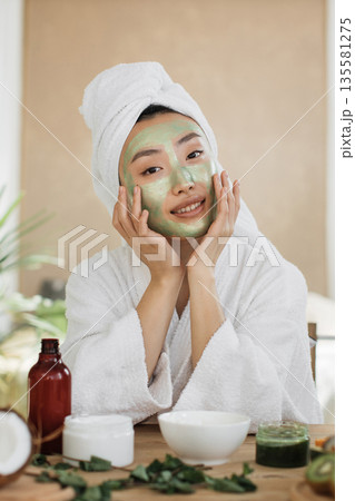 A young woman with a towel wrapped around her head applies a green facial mask while wearing a white bathrobe, surrounded by skincare products and natural ingredients A young woman with a towel wrapped around her head applies a green facial mask while wearing a white bathrobe, surrounded by skincare products and natural ingredients 135581275