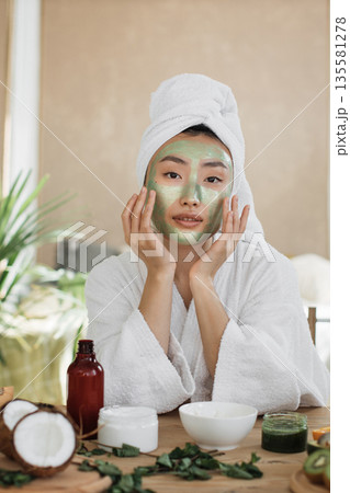 A young Asian woman with a towel wrapped around her head applies a green clay mask to her face while wearing a white bathrobe, surrounded by natural beauty products 135581278
