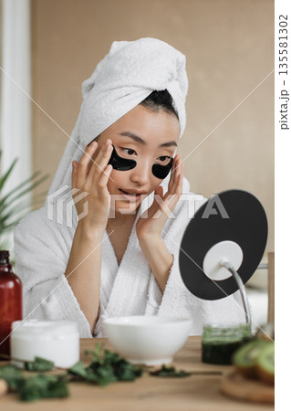A young Asian woman in a bathrobe and towel applies black under-eye patches while looking in a mirror, surrounded by skincare products and greenery A young Asian woman in a bathrobe and towel applies black under-eye patches while looking in a mirror, surrounded by skincare products and greenery 135581302