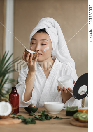 A young woman with a towel wrapped around her head and a white robe applies cream to her face while smelling a piece of coconut 135581336