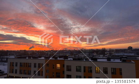 Bright colorful sunset over a city skyline with buildings and industrial pipes emitting smoke and a snow-covered landscape. Nature photography. 135581574