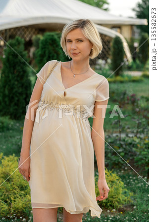 Pregnant woman in beige dress standing in garden during sunny day near tents and greenery 135582673