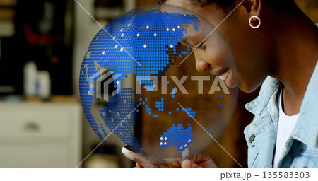Holding smartphone, woman in jacket showing blue globe overlay above phone in kitchen, copy space 135583303
