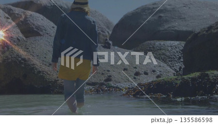 Wading woman wearing straw hat and denim jacket through tidal pool with seaweed on boulders 135586598