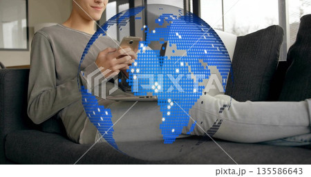 Reclining casually dressed man using smartphone, laptop in living room, with digital globe overlay 135586643