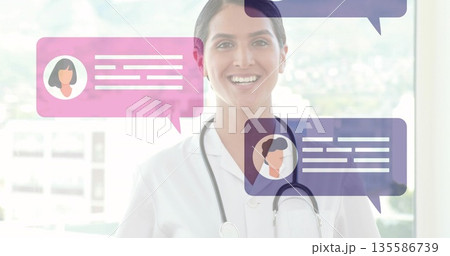 Smiling female physician standing in clinic sporting white coat and stethoscope with chat bubbles 135586739