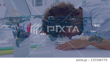 Leaning fatigued office worker pressing head onto desk at office, with laptop and world map overlay 135586779