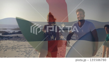 Walking friends wearing swimwear carrying surfboards at sandy shoreline, with ocean waves and rocks 135586780