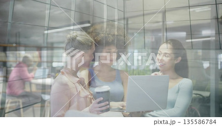 Collaborating three women working on laptop in office lounge, with glass partition and coffee cup Collaborating three women working on laptop in office lounge, with glass partition and coffee cup 135586784