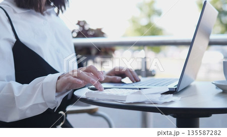 A business woman types on her laptop while sitting at a round table outside a cafe. She appears focused and engaged in her work during a sunny day with greenery in the background. A business woman types on her laptop while sitting at a round table outside a cafe. She appears focused and engaged in her work during a sunny day with greenery in the background. 135587282