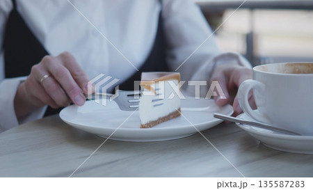 A woman in a white shirt delicately holds a fork to enjoy a slice of dessert on a white plate beside her cup of coffee. She appears focused on her treat while seated at a cafe. A woman in a white shirt delicately holds a fork to enjoy a slice of dessert on a white plate beside her cup of coffee. She appears focused on her treat while seated at a cafe. 135587283