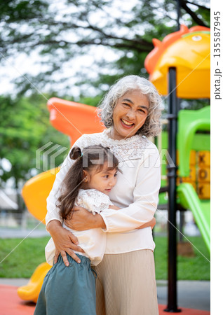 Old woman grandmother and kid girl granddaughter standing holding or hugging in park's playground. 135587945