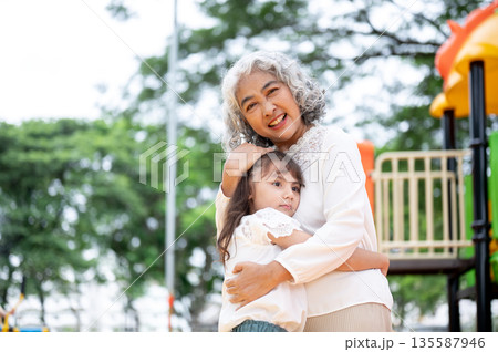 Old woman grandmother head patting kid girl granddaughter as standing hugging in park's playground. Old woman grandmother head patting kid girl granddaughter as standing hugging in park's playground. 135587946