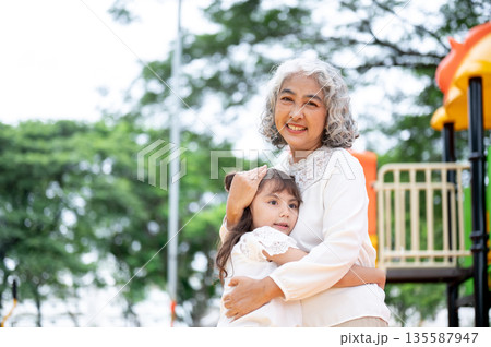 Old woman grandmother patting head kid girl granddaughter as standing hugging in park's playground. Old woman grandmother patting head kid girl granddaughter as standing hugging in park's playground. 135587947