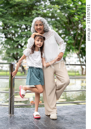 Old woman grandmother holding hand standing with kid girl granddaughter at railing around the lake. 135587951