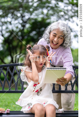 Kid girl granddaughter making funny face imitating a drawing in notebook holding by old woman granny Kid girl granddaughter making funny face imitating a drawing in notebook holding by old woman granny 135588009