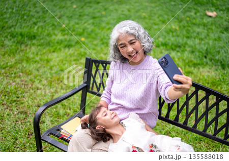 Old woman grandma taking picture on phone with kid girl granddaughter laying on lap at park's bench. Old woman grandma taking picture on phone with kid girl granddaughter laying on lap at park's bench. 135588018