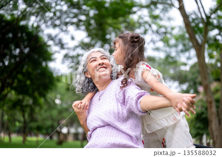 Old woman grandmother smiling while playing with a kid girl granddaughter behind her back in park. Old woman grandmother smiling while playing with a kid girl granddaughter behind her back in park. 135588032