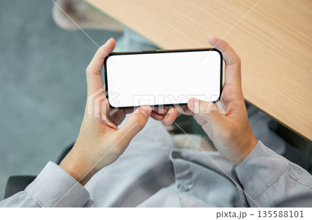 Blank screen smartphone holding horizontally with both hand while sitting at wooden table in a cafe. 135588101