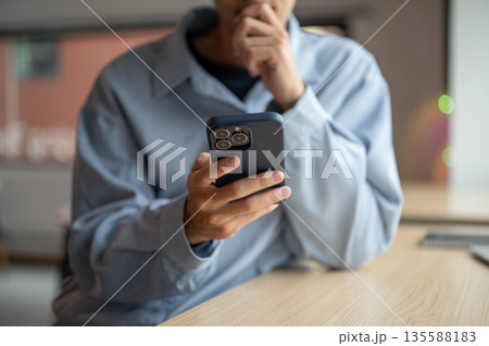 Close up of a man looking at smartphone with hand holding chin while sitting at wooden table in cafe Close up of a man looking at smartphone with hand holding chin while sitting at wooden table in cafe 135588183