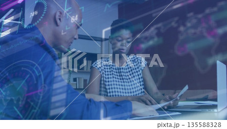 Reviewing documents and typing on laptop two colleagues at office desk, with holographic charts Reviewing documents and typing on laptop two colleagues at office desk, with holographic charts 135588328