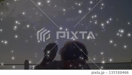Sitting couple on wooden railing facing ocean horizon at pier dusk with sparkling stars Sitting couple on wooden railing facing ocean horizon at pier dusk with sparkling stars 135588466