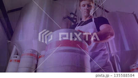 Brewery worker wearing white apron lifting keg on brewery floor, with fermentation tanks, pipes Brewery worker wearing white apron lifting keg on brewery floor, with fermentation tanks, pipes 135588584