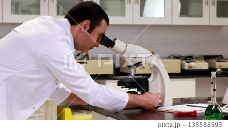 Adjusting microscope focus knob, technician wearing lab coat at lab bench with flask, copy space 135588593