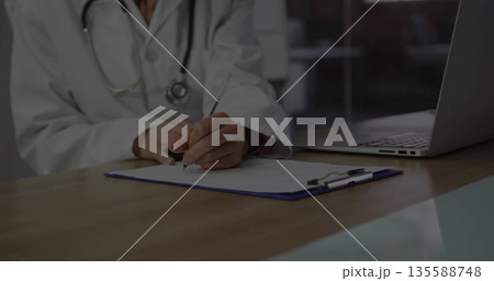 Sitting doctor in lab coat writing on clipboard in clinic, with stethoscope and laptop, copy space Sitting doctor in lab coat writing on clipboard in clinic, with stethoscope and laptop, copy space 135588748