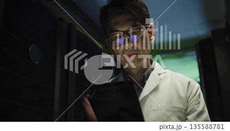 Analyzing tablet data, Korean man in lab coat standing between server racks, showing digital charts Analyzing tablet data, Korean man in lab coat standing between server racks, showing digital charts 135588781
