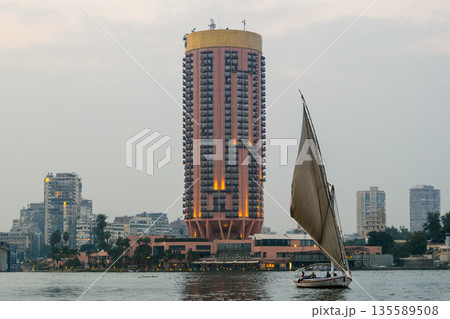 A traditional felucca sailing on the Nile River in Cairo. The boat has a large sail and is surrounded by modern buildings along the riverbank. A traditional felucca sailing on the Nile River in Cairo. The boat has a large sail and is surrounded by modern buildings along the riverbank. 135589508