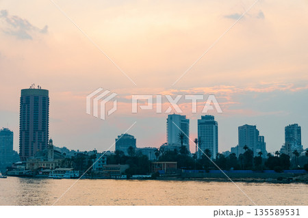 Sunset over the Nile River in Cairo. A tall building stands by the riverbank, with the sun setting behind it, casting warm colors in the sky. Sunset over the Nile River in Cairo. A tall building stands by the riverbank, with the sun setting behind it, casting warm colors in the sky. 135589531