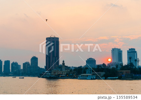Sunset over the Nile River in Cairo. A tall building stands by the riverbank, with the sun setting behind it, casting warm colors in the sky. Sunset over the Nile River in Cairo. A tall building stands by the riverbank, with the sun setting behind it, casting warm colors in the sky. 135589534