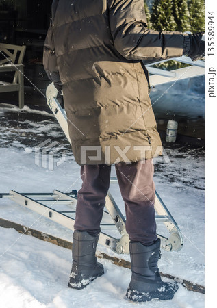Man in winter clothes carrying a folding ladder in the snow. Man in winter clothes carrying a folding ladder in the snow. 135589944