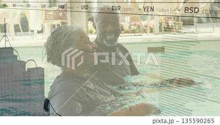 Senior woman, African American man chatting in resort pool on tiled deck, wearing dark swim shirts 135590265