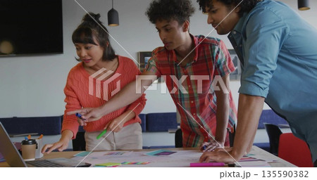 Pointing and discussing printed layouts around modern meeting table with laptop, sticky notes 135590382