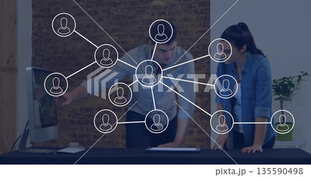 Leaning colleagues using desktop PC and tablet on desk in office, plant, network icons overlaying 135590498