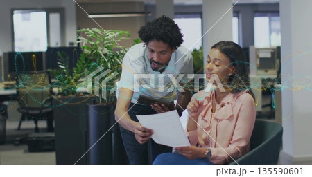 Office professionals reviewing printed document in open-plan office, with tablet by potted plant 135590601