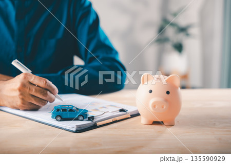 Man signs car finance contract near pink piggy bank as money savings for auto insurance and lease documents, using price management to save for buying vehicle on wooden office desk Man signs car finance contract near pink piggy bank as money savings for auto insurance and lease documents, using price management to save for buying vehicle on wooden office desk 135590929