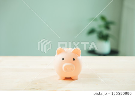 Pink piggy bank for money saving goals sits on wooden table, financial investment concept for personal finance, background showing blurred houseplant and minimalist home interior Pink piggy bank for money saving goals sits on wooden table, financial investment concept for personal finance, background showing blurred houseplant and minimalist home interior 135590990